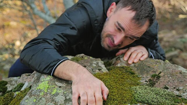 A man is seen gripping a mossy rock surface, showcasing determination as they navigate through a rugged, natural environment. The soft-focus background highlights the surrounding foliage. Close-up