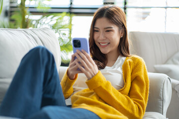 Happy young asian woman using mobile apps in living room,Smiling young asian female using smartphone sitting on couch at home.Messaging,Online Shopping,celebrate success,chatting online,Social Media