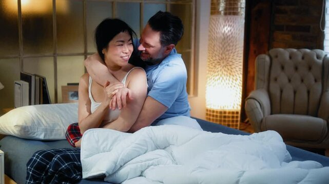 Happy multiracial couple in pajamas hugging and cuddling in bed after waking up in the morning at home in the bedroom