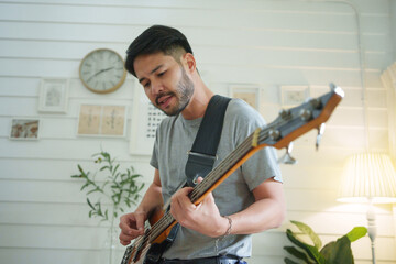 Asian man enjoys playing bass guitar, creating a relaxing. Concentrated Male Bassist practicing his...