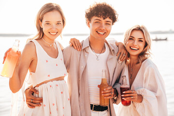 Young stylish friends posing at the seaside. Fashion man and two cute female dressed in casual summer clothes. Smiling models having fun. Cheerful women and guy outdoors, Hold and drink lemonade