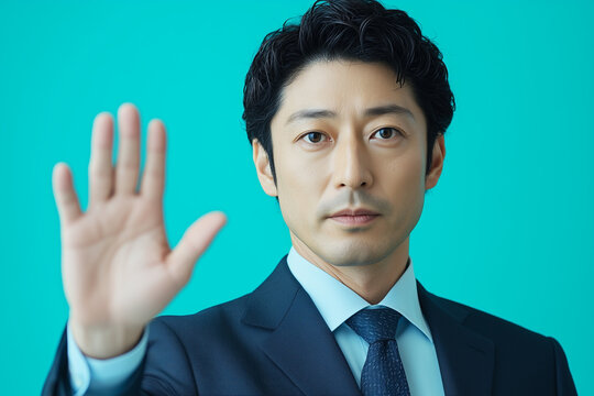A Japanese man in a suit, with his right hand raised as if saying "no," facing the camera directly.