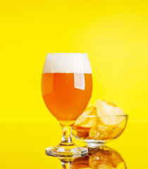 A glass of cold beer alongside a pile of crispy potato chips on a vibrant yellow background