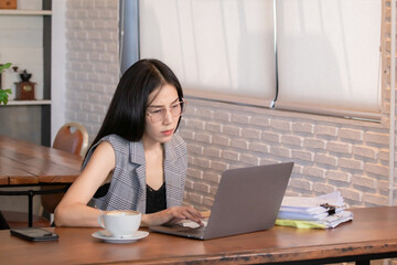 Young Asian female sitting in coffee shop workplace using laptop with stacks of bills or invoices, digital nomad working everywhere remotely from a cafe, businesswoman lifestyles working concept