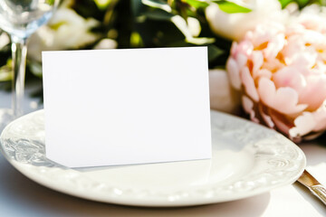 Close-up of white blank guest seating card mockup on plate. Elegant wedding table setting with peonies