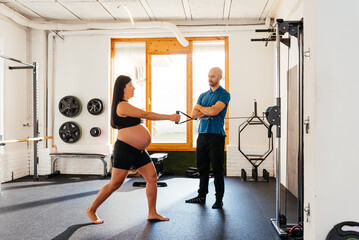 Fototapeta premium Pregnant Chinese Woman Exercising With Resistance Cables Under the Guidance of a Trainer in a Gym Setting