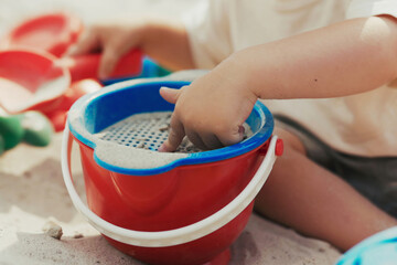 Boy in the backyard plays in the sandbox, pouring sand from a shovel into a bucket, building sandcastles on his own.