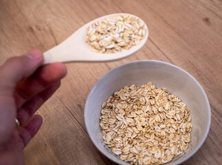 A hand holds a wooden spoon filled with raw rolled oats with a bowl of oats blurred in the background. The scene is set on a wooden surface, creating a natural and rustic feel.