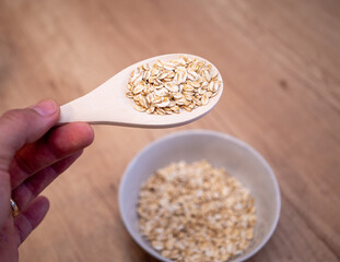 A hand holds a wooden spoon filled with raw rolled oats with a bowl of oats blurred in the background. The scene is set on a wooden surface, creating a natural and rustic feel.