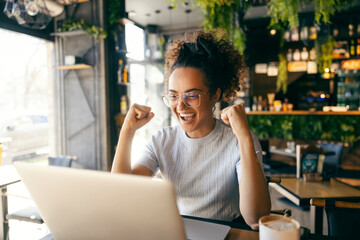 Cheerful female entrepreneur sitting in cafe, looking at laptop and cheering for winning price.