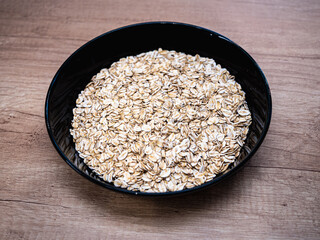A black bowl filled with raw rolled oats sits on a wooden countertop. The simple composition highlights the natural texture and color of oats, commonly used for healthy meals.