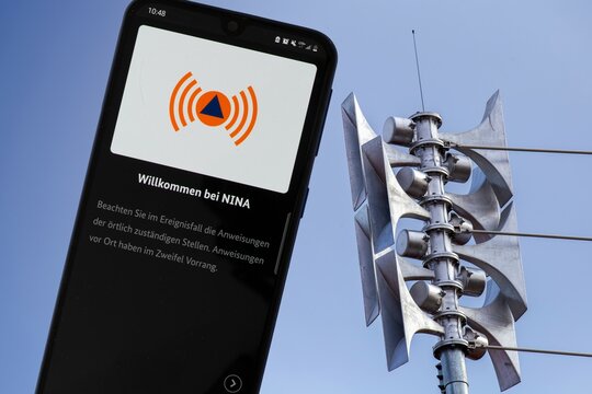 (Fotomontage) Nahaufnahme einer Sirene an einer Schule, daneben die ge&ouml;ffnete Warn-App NINA auf einem Smartphone