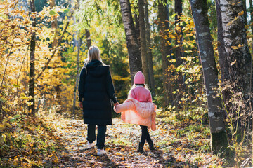 Fototapeta premium Mother and daughter are walking in an autumn park filled with sun rays, photo from the back.