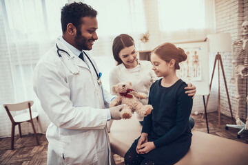 Indian doctor seeing patients in office. Doctor is giving mother and daughter teddy bear.