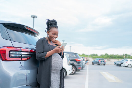 Donna africana incinta con capelli intrecciati sorride mentre si appoggia a un'auto in un parcheggio all'aperto, indossando un vestito grigio e camicia nera, guardando felice il suo smartphone sotto u