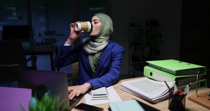 Young woman office worker in hijab sits at desk and sips hot coffee. Tired secretary tries to finish urgent assignment at thunderstorm in evening