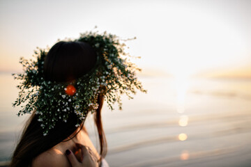 A girl in a wreath of wildflowers stands by the sea at sunset