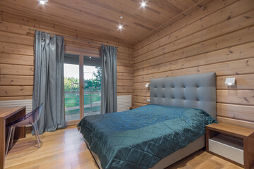 A delightful bedroom interior in a luxurious mansion made of light wooden beams .Balcony door with a view of the lawn. Stylish furniture.