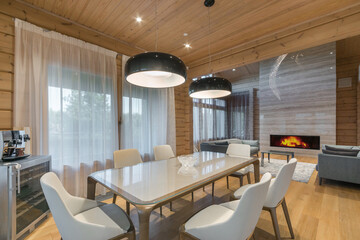 Stylish interior of the living room in a luxurious wooden mansion. A dining table with white chairs, upholstered sofas and a fireplace.