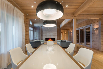 A long dining table with white chairs in the interior of the living room of a luxurious wooden mansion. Fireplace and soft sofas.