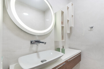 Bathroom with white interior decor. Hanging shelves, a round mirror with illumination above the sink.