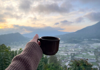 A woman’s hand holding a cup of coffee with beautiful morning view of mountains 
