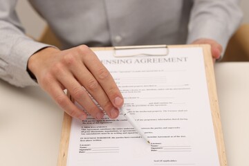 Man with pen pointing at licensing agreement document at white table indoors, closeup