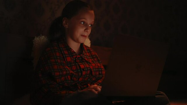 Portrait of happy pretty child girl 10 yo with joyful expression using laptop sitting on bed in dark bedroom at night, warm lighting creating cozy and intimate atmosphere. Shooting in slow motion.