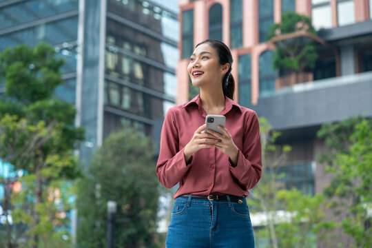 Successful asian business woman using phone outdoors.Happy businesswoman holding cellphone using smartphone or mobile phone standing or walking on city urban street,5g communicating with a colleague.
