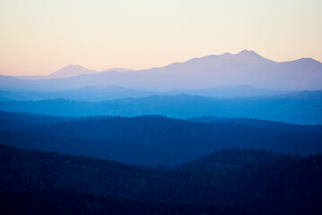 Obraz premium Serene view of rolling mountains in shades of blue and green, with distant peaks under soft, pastel sky, creating peaceful and tranquil landscape.