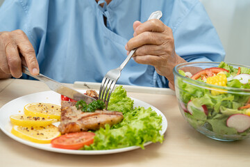Asian senior woman patient eating pork chop stake and vegetable salad for healthy food in hospital.