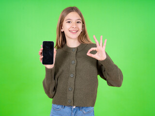 beautiful caucasian girl wearing green sweater holding in hands cell showing ok-sign