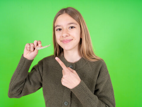 beautiful caucasian girl wearing green sweater holding an invisible aligner and pointing at it. Dental healthcare and confidence concept.