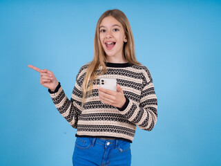 Obraz premium Astonished beautiful caucasian girl wearing striped sweater holding her telephone and pointing with finger aside at empty copy space