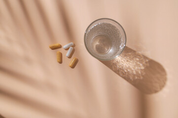 Glass of water, vitamins, capsules, dietary supplements, probiotics, tablets on beige background with beautiful long shadows from sun. Nutraceuticals, health care. Medical pharmaceutical background