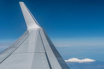 The wing of a plane in flight as it flies over the snow-capped mountains on the border between...