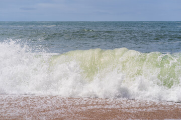 Fototapeta premium Powerful wave crashes onto the shore, raising white foam on golden sand, seascape on a cloudy day.