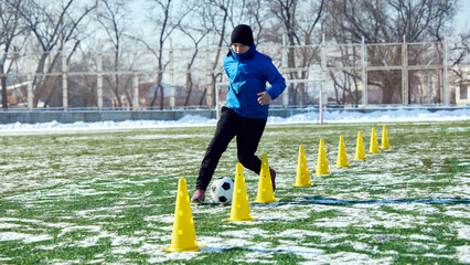 Football player dribbling soccer ball through yellow cones on outdoor field, improving control and coordination. Concept of sport, school, childhood, training, skill development, active lifestyle.
