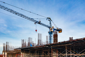 A construction site with a blue crane towering over reinforced concrete columns, steel rebar, and...