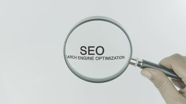 SEO (Search Engine Optimization) text or word highlighted by a magnifying glass in a hand on a white background, symbolizing focus on improving online visibility, rankings, and website performance