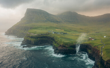 Aerial view of dramatic cliffs and a picturesque waterfall cascading into the ocean near a remote village, Gasadalur, Vagar, Faroe Islands.
