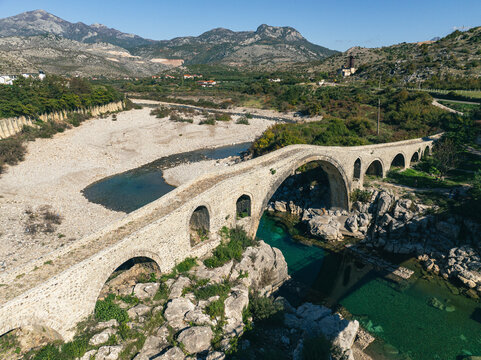 Aerial view of Mesi Bridge over a scenic river surrounded by historic mountains and lush greenery, Shkoder, Shkoder, Albania.