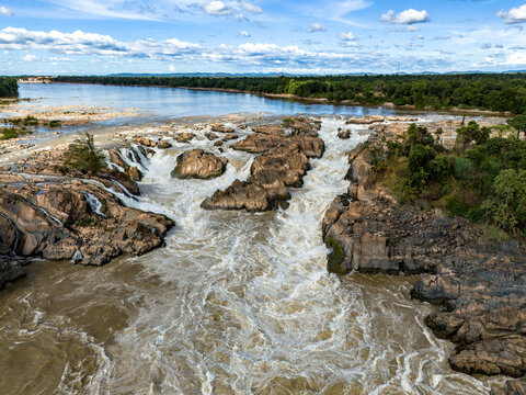 Aerial view of majestic waterfall cascading over rocks amidst lush greenery and flowing river, Khong District, Champasak, Laos.