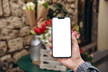 Mockup woman's hand holding using smart phone with white blank screen POV at flower street shop, mobile app design and advertising, online marketing