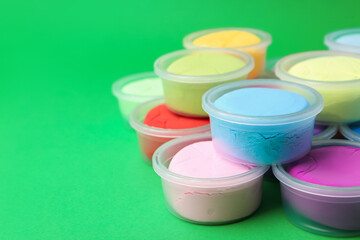 Different colorful modeling clay in plastic jars on green background, closeup. Space for text
