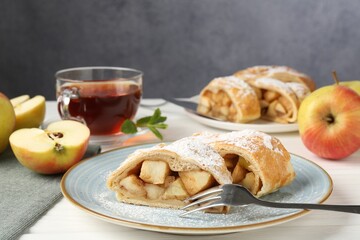 Pieces of tasty apple strudel with powdered sugar and fork on white wooden table against grey background, closeup
