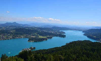Naklejka premium Aerial view of a historic village and church with a tall spire on the shores of Wörthersee, a stunning blue lake in the Austrian Alps. Green forests and mountains. European destination. Nature. 