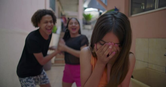 Young girl hiding face in hands while being mocked by friends in suburban alleyway, highlighting themes of bullying, peer pressure, and emotional isolation