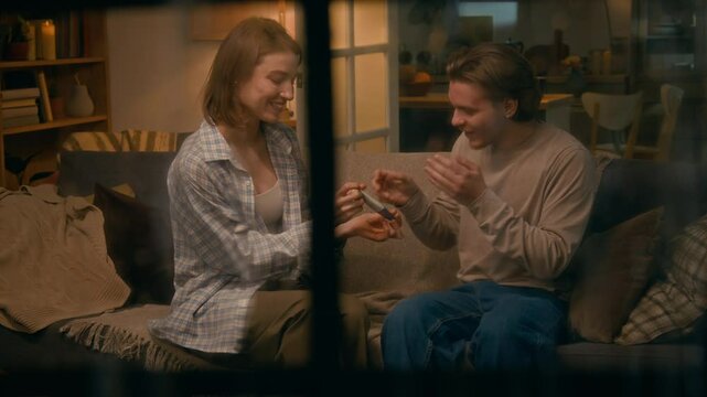 Medium shot, observed through window on rainy evening, of cheeky young Caucasian wife sitting on couch, taking out pregnancy test with two stripes from behind back and giving to husband