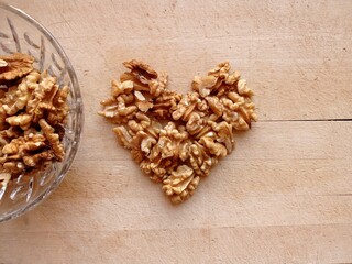 Top view of some shelled walnuts arranged in the shape of a heart on a wooden board. A few nuts are in the small glass bowl next to the heart.